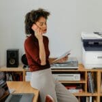 Imprimante multifonction couleur Xerox® C325 dans un environnement de bureau à domicile Une femme assise à un bureau en train de passer un appel à côté d'une imprimante Xerox® C325